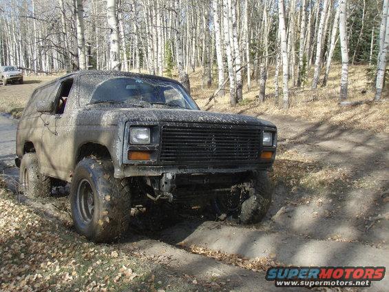 Mud-covered SUV with a black grille and square headlights, driving on a dirt path in a forest. Another vehicle is visible in the background