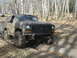 1985 Ford Bronco - An off-road SUV navigates a muddy trail through a wooded area, with another vehicle in the distance.