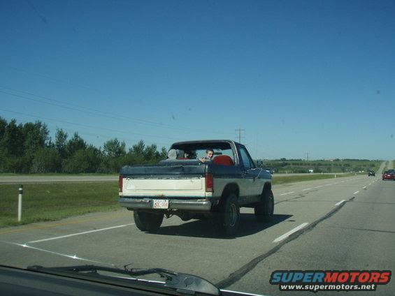 Rear view of a blue and white Ford Bronco driving on a highway. Two people are in the cab