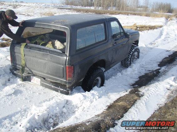 Dark gray Ford Bronco stuck in deep snow, person pushing on rear hatch. License plate visible