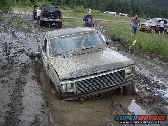 Mud-covered pickup truck stuck in a muddy rut, front view. Other vehicles and people are visible in the background
