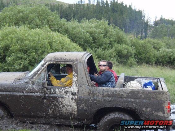 Mud-covered 1980s Ford Bronco stuck in mud with four people inside, driving through a wooded area