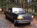 1995 Ford F-150 - A black 1995 Ford F-150 pickup truck is parked on a road covered in fallen leaves, surrounded by trees.