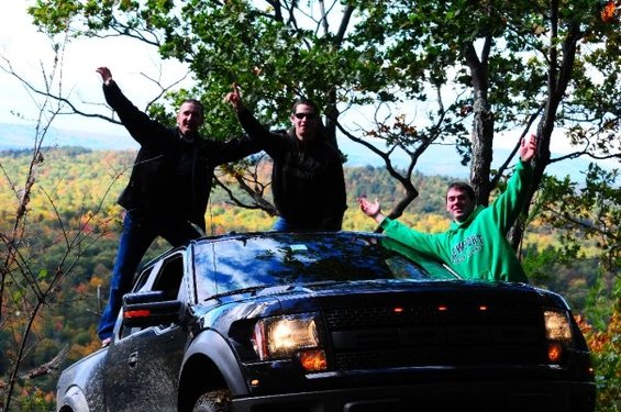 Black Ford F-150 Raptor with three guys on top, arms raised, in wooded area