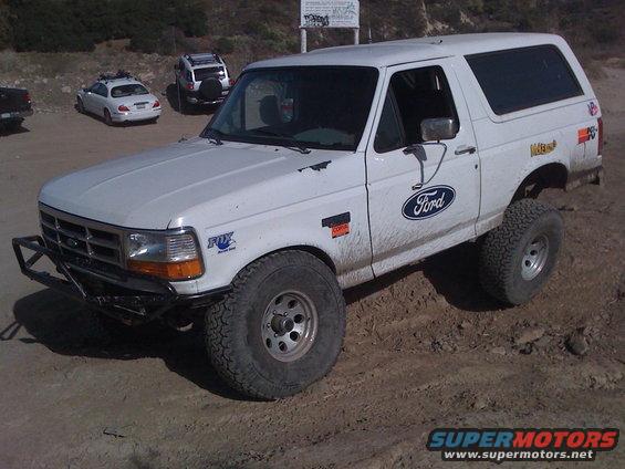 White Ford Bronco with aftermarket bumper and off-road tires parked on dirt. Other cars are in the background