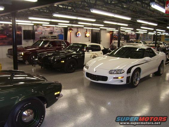 Several cars parked inside a garage, including a white Camaro and a black Camaro