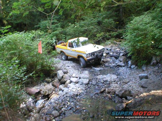 1978 Ford Bronco navigating a rocky stream bed with trees in the background