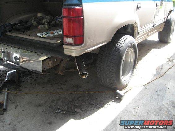 SUV rear bumper, taillight, and tire. Chrome bumper, red taillight, exhaust pipe. License plate visible