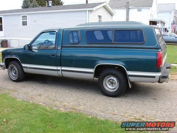 side view of a green pickup truck with a camper shell, parked on a cracked asphalt driveway