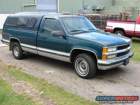 Green pickup truck with a silver stripe and a camper shell parked on cracked asphalt. Another red and white truck is visible in the background