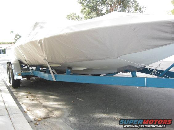 Boat covered with a grey tarp on a blue trailer, parked on asphalt next to a curb