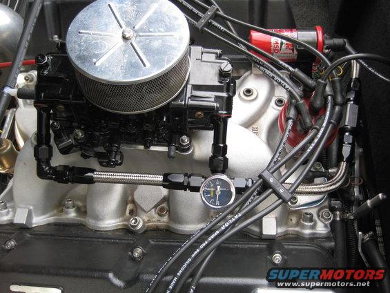 Close-up of a V8 engine's intake manifold, carburetor, air cleaner, fuel lines, and spark plug wires. A small fuel pressure gauge is visible