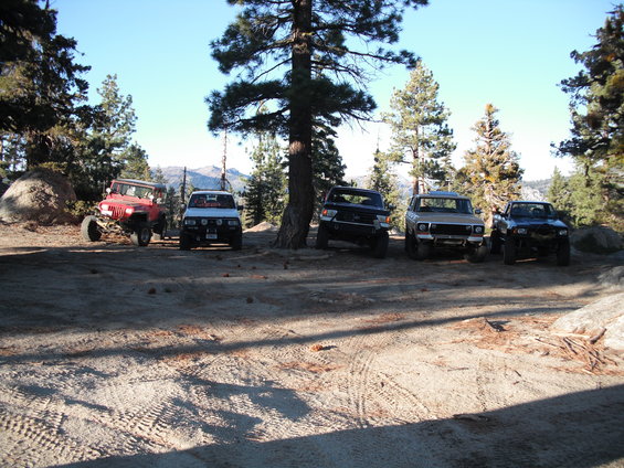 Several off-road vehicles parked on a dirt path near trees, with mountains in the background