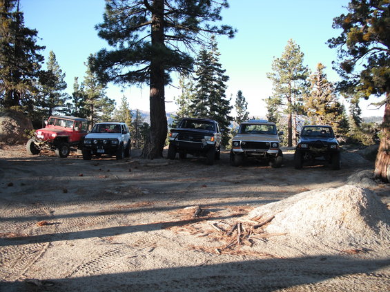 Five off-road vehicles parked in a wooded area with a pile of sand in the foreground