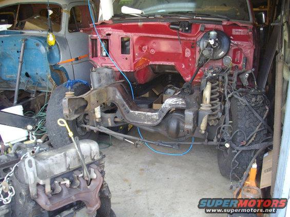 Disassembled vehicle chassis with engine and another vehicle body in background