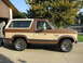 1986 Ford Bronco - This two-tone Ford Bronco is parked outside a garage with trees in the background.