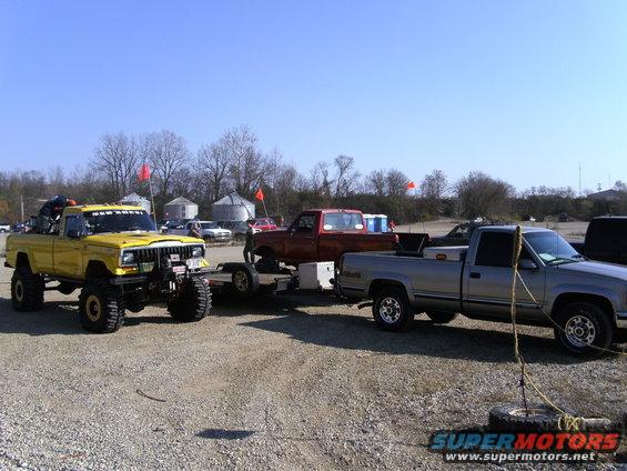 Yellow Jeep Comanche pickup with big tires, towing a trailer. A red truck and a silver truck are behind it