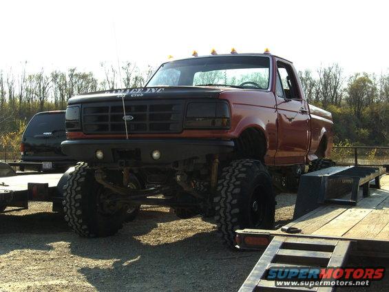 Red lifted Ford pickup truck with black grille and hood decal, parked on gravel next to a trailer