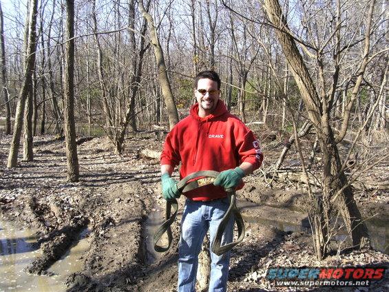 Man in red hoodie and green gloves holds a tow strap in a muddy, wooded area. Watermark reads SUPERMOTORS.NET