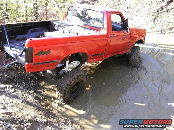 Red pickup truck with lifted suspension and large tires driving through a muddy puddle