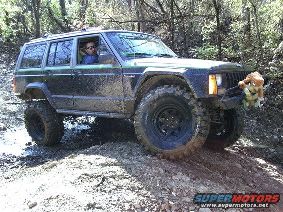 Black Jeep Cherokee XJ with green trim, lifted suspension, and large tires on a muddy trail