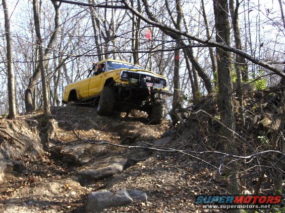 Yellow Jeep Comanche pickup truck with large tires climbing a rocky, wooded trail