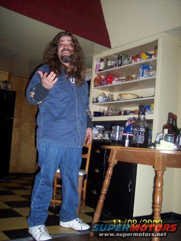 Man with long curly hair and beard wearing a blue shirt and jeans, standing in a room with shelves of items and a table
