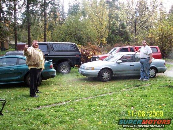 Light blue sedan parked on grass with two men nearby; red pickup truck and black SUV behind it