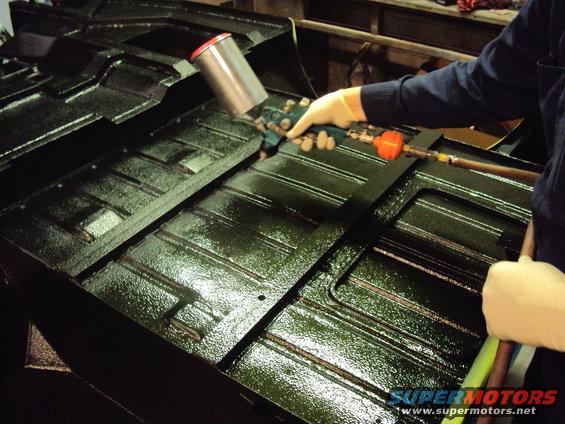 Person spraying dark green protective coating onto a car's floor pan with a spray gun