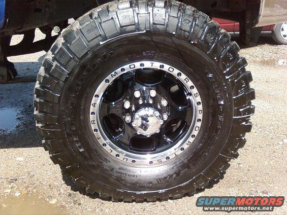 Truck tire with aggressive tread and black wheel with chrome center cap. A blue truck frame is visible above