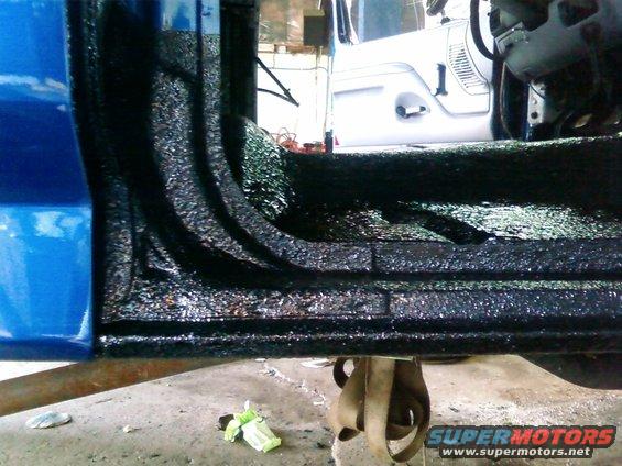 Close-up of a blue truck's interior floor with black spray-on bed liner. A white Jeep is visible in the background
