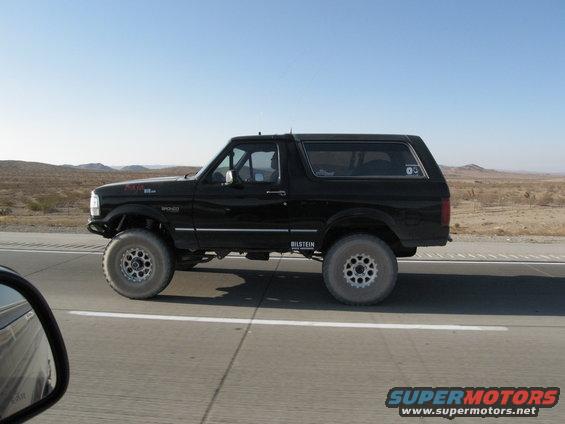 Black Ford Bronco with aftermarket wheels and suspension driving on a highway. Bilstein shocks visible