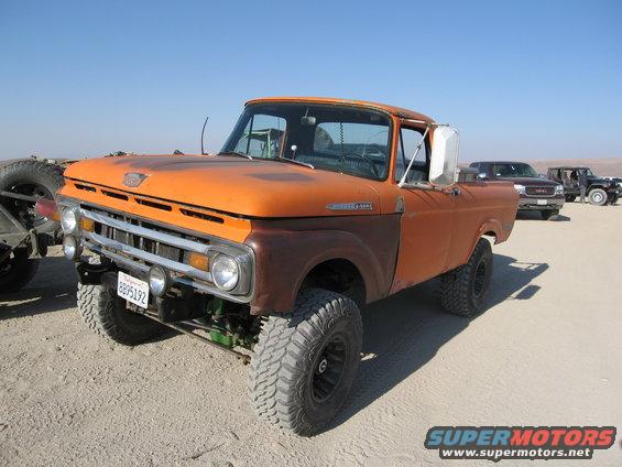 Orange pickup truck with lifted suspension and off-road tires parked on dirt. Other trucks are in the background
