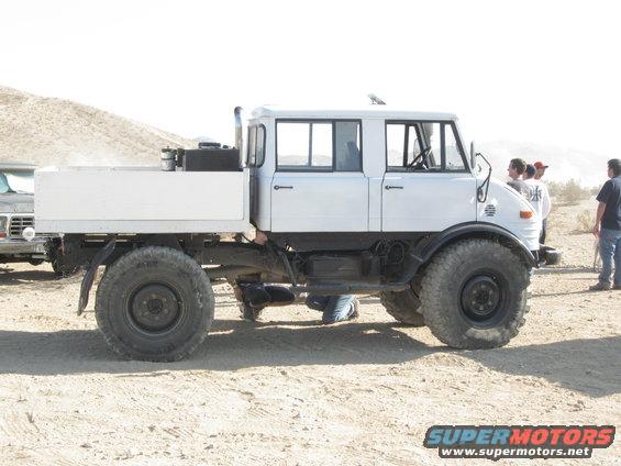 White flatbed truck with a crew cab, large tires, and a flatbed cargo area, parked on a dirt surface