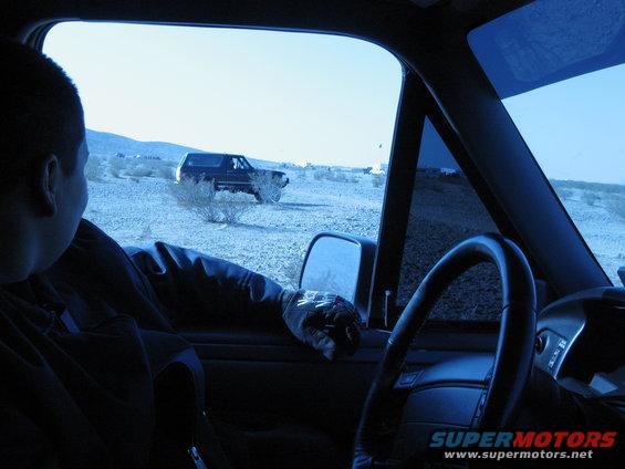 View from inside a vehicle looking out at a dark blue 1987 Ford Bronco parked in a desert
