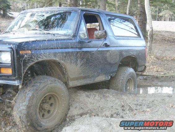 Mud-splattered dark blue 1980s Ford Bronco stuck in mud, front passenger side wheel deep in muck