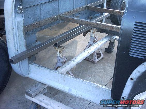 30964780094_large.jpg 10-15-08:  I also started to lay out the rear crossmember that I'm fabricating to support the rear floor section. The crossmember piece is the bottom tube sitting on the jack stands. Tomorrow I'm going to cut out the down tubes for the crossmember. I'm building it out of 2"x2" - 1/8" wall tubing. So I shouldn't have to worry about falling thru the floor!  Nothing like a little overkill!! There is a channel in the bottom of the 78 rear floor section that the 2" tube fits perfect in. I'm going to weld the crossmember to the rocker plates and then weld the floor to the crossmember.