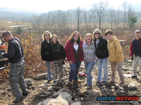guy working on vehicle, group of women standing behind him, off-road terrain