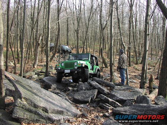 Green Jeep Wrangler on rocky terrain in a forest, with another black SUV behind it