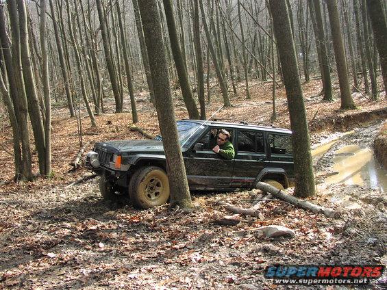 Dark green Jeep Cherokee XJ navigating a muddy forest trail, driver visible