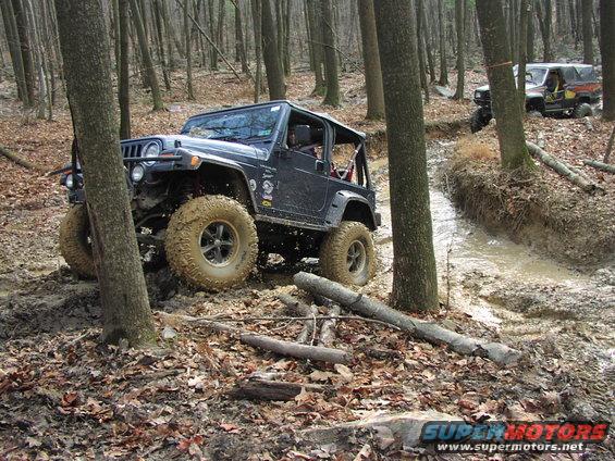 Black Jeep Wrangler navigating a muddy forest path with another Jeep visible in the background