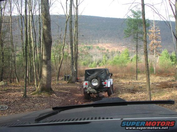 Jeep Wrangler driving on a dirt path in a wooded area, another vehicle is visible in the background