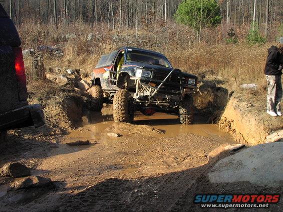 Off-road SUV with large tires driving through a mud pit, another vehicle visible on the left