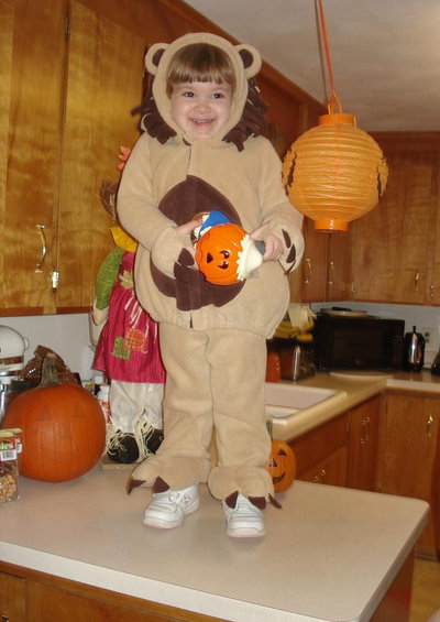 Child in lion costume stands on kitchen counter holding a pumpkin candy bucket. A large pumpkin is to the left