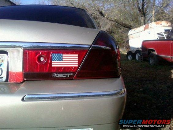 Silver sedan's rear tail light with an American flag sticker and SCT emblem, parked outdoors