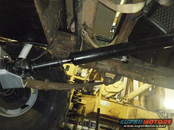 Underside view of a vehicle's drivetrain and suspension components, with a yellow piece of heavy equipment in the background