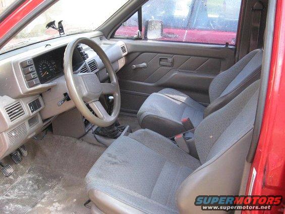 Interior shot of a red vehicle, showing the dashboard, steering wheel, and front seats. A red truck is visible in the side mirror
