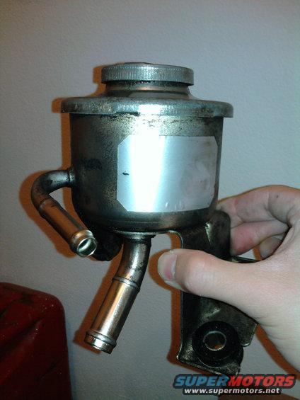 Hand holding a metal power steering fluid reservoir with two hose barbs and a cap