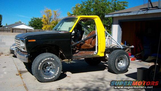 Modified pickup truck with black front end and yellow cab, roll cage, and aftermarket wheels. Visible text on tires reads 'Mud King XT'