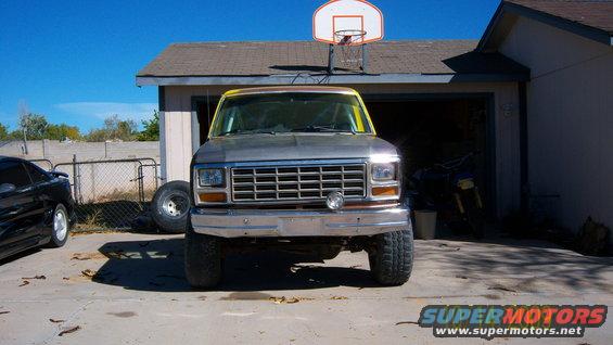 Front view of a lifted Ford F-Series truck with a yellow top, parked in a driveway. A black car is on the left